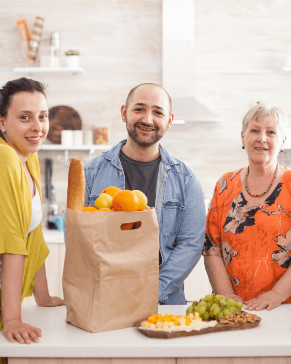 about second img Four people around a kitchen counter with a grocery bag and fresh fruit.